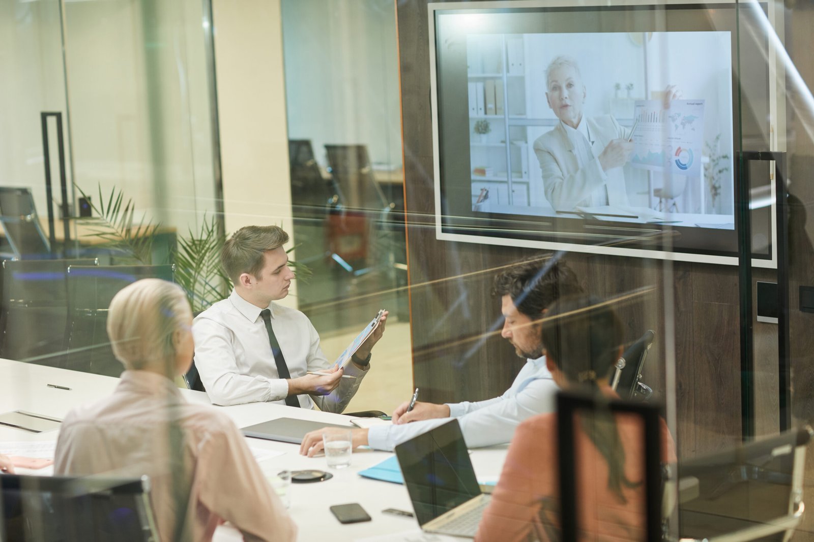Group of business people sitting at the table and discussing new project with mature businesswoman during online conference behind the glass wall
